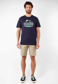 T-shirt en coton bleu marine avec un graphisme "OXBOW" en couleurs claires, associé à un short cargo beige. Baskets noires avec semelles blanches.