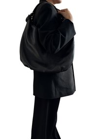 Osoba nosící černé sako a kalhoty, přenášející velkou černou texturovanou koženou tašku přes pravé rameno před bílým pozadím.