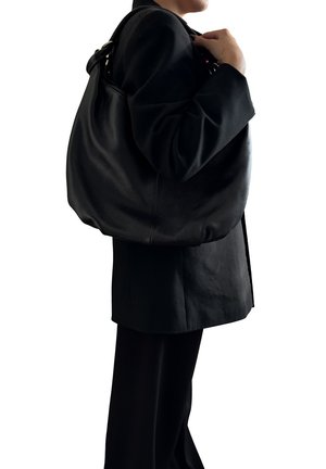 Persona che indossa un blazer nero e pantaloni, con una grande borsa a tracolla in pelle nera texturizzata sulla spalla destra, su sfondo bianco.