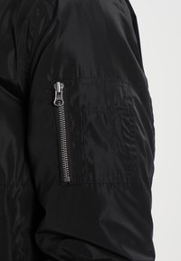 Schwarze Bomberjacke mit gesteppter Textur, ausgestattet mit einer seitlichen Reißverschlusstasche und dezentem Glanz. Der Stoff ist glatt und leicht.