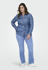 Denim srajca s vrvico za prilagajanje v pasu, dolgimi rokavi, dvema žepoma na prsih in svetlo modro barvo. V kombinaciji s svetlo modrimi kavbojkami in belimi čevlji.