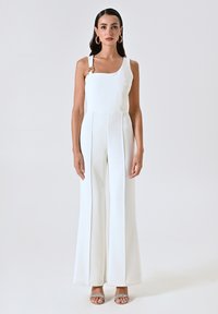 Jumpsuit blanc avec un buste ajusté, des pantalons larges et une seule bretelle avec un accent doré. Texture lisse avec des détails de couture.