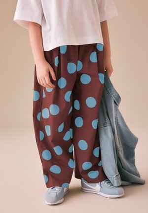 Barn iført hvid skjorte, store brune bukser med store blå polkaprikker, lyseblå sneakers, der holder en lyseblå denimjakke.