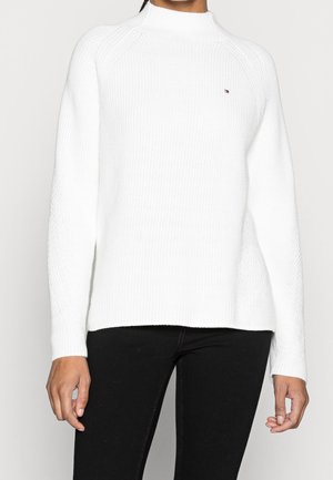 Personne portant un pull en maille côtelée blanc avec un petit logo sur la poitrine et un pantalon noir, se tenant devant un fond uni.