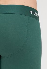 Leggings de culoare verde închis cu o textură netedă, având un design ajustat și un talie etichetată "HOLLISTER." Detaliile cusăturii sunt minimale.