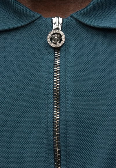 Žalsvai mėlynas užsegamas drabužis su tekstūruota paviršiaus struktūra. Yra sidabrinės spalvos užtrauktukas ir apvalus užtrauktuko laikiklis su liūto emblemą ir graikiško raktų rašto ornamentą.