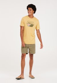 Geel katoenen t-shirt met een logo, gecombineerd met gestructureerde groene shorts en bruine teenslippers. Model staat in een casual houding.