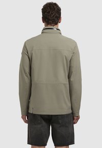 Jachetă de un verde măsliniu deschis, cu guler înalt, textură mată și detalii pe cusătura din spate; combinată cu pantaloni scurți din denim închis.