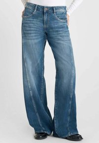 Jean en denim bleu à jambes larges avec une taille haute, présentant un délavage subtil, un design classique à cinq poches et des coutures contrastantes le long des coutures.