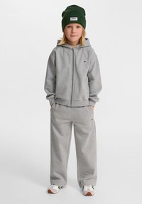 Grå zip-up hoodie och matchande sweatpants i mjukt material, matchat med en grön mössa. Har logotyp på bröstet och byxorna.