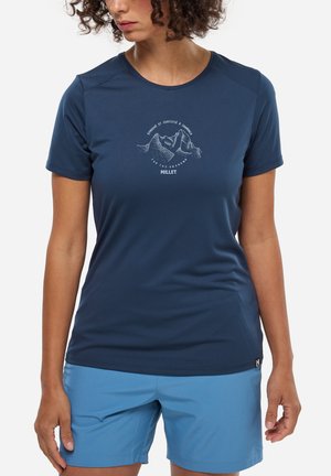 Femme portant un t-shirt à manches courtes bleu marine avec un motif de montagne et un short bleu clair, debout devant un fond blanc uni.