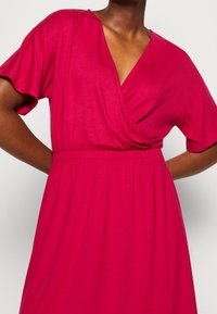 Robe enveloppante fuchsia avec des manches courtes, taille élastique et tissu doux. Présente un décolleté en V profond et une silhouette fluide.