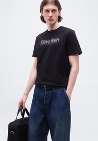 Νέος άνδρας που φοράει μαύρο T-shirt Calvin Klein και μπλε τζιν, κρατώντας μια μαύρη τσάντα με το ένα χέρι στην τσέπη του.