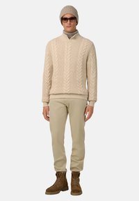 Beige kabelstickad tröja med hög krage, beige byxor och bruna snörskor. Tillbehören inkluderar beige mössa och solglasögon.