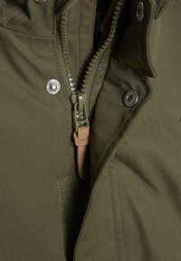 Chaqueta verde oliva con textura de espiga, cierre de cremallera con acento de cuero y detalles de botones a presión para un ajuste seguro.