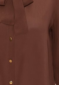 Blouse marron à manches longues avec des boutons ronds dorés et un détail noué au col.