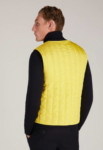 Gelbe gesteppte Weste mit hohem Kragen, getragen über einem dunkelblauen Pullover. Die Weste hat vertikale Nähte und eine glatte Textur.