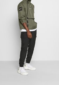 Chaqueta ligera verde oliva con un panel de acento negro, combinada con pantalones cargo negros y zapatillas blancas con detalles en dorado.