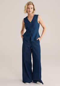 Tumši zilā džinsu veste un plašas bikses. Vestē ir V-veida kakla izgriezums, bez piedurknēm un priekšējie pogas; biksēm ir brīvs piegriezums un sānu šuves.