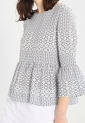 Femme portant un chemisier blanc avec des motifs géométriques noirs et des manches évasées trois quarts, associé à un pantalon blanc.