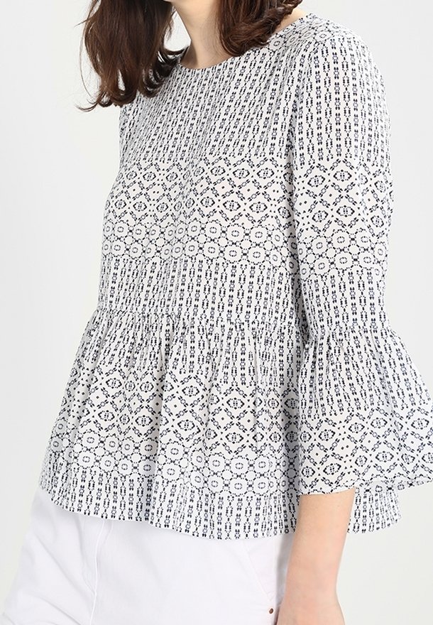 Femme portant un chemisier blanc avec des motifs géométriques noirs et des manches évasées trois quarts, associé à un pantalon blanc.