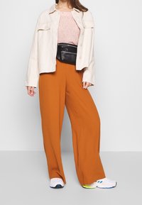 Beżowa kurtka cropsowa założona na różowy sweter, zestawiona z pomarańczowymi szerokimi spodniami i białymi trampkami. W zestawie znajduje się czarna saszetka na pas.