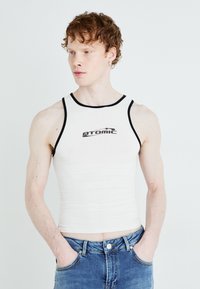 Vit ärmlös tanktopp med svart kant, med en centrerad "ATOMIC" logotyp. Bärs med blå denimjeans som har en figurnära design.