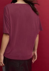 Haut ample à manches courtes en tissu bordeaux profond, texture lisse, épaules tombantes et encolure large, présenté sur un fond rouge.