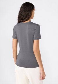 Vrouw met lang, donker haar draagt een strakke, kort-sleeve grijze shirt en off-white broek, staand tegen een effen witte achtergrond met haar rug naar de kijker toegekeerd.