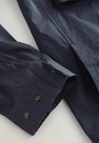 Cuff di una camicia a maniche lunghe blu scuro con due bottoni a pressione neri e oro su uno sfondo chiaro.