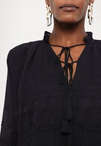 Blusa preta bordada com padrões florais, decote com babados e laços com franjas. Apresenta um tecido texturizado e destacados acentos pretos.