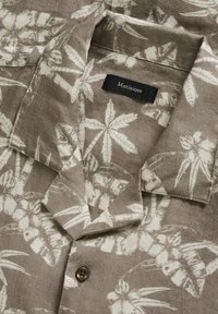 Camisa de manga corta en lino marrón claro con un patrón de hojas de palma blancas, cuello con botones y una pequeña etiqueta que dice "Matinique."