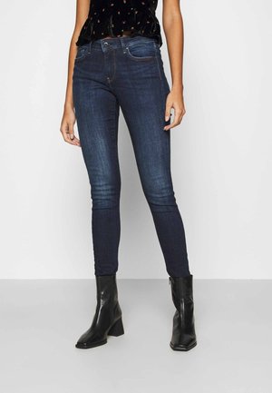 Jeans skinny de color azul oscuro, hechos de denim, con una cintura de altura media y un ligero desgastado a lo largo de las piernas, combinados con botas negras hasta el tobillo.