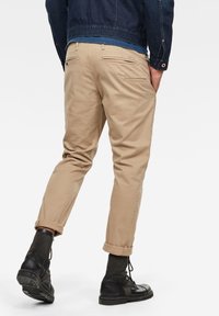 Tan chinos with a tapered fit, rolled cuffs, and rear pockets. Paired with black lace-up boots. Denim jacket visible on the upper body.