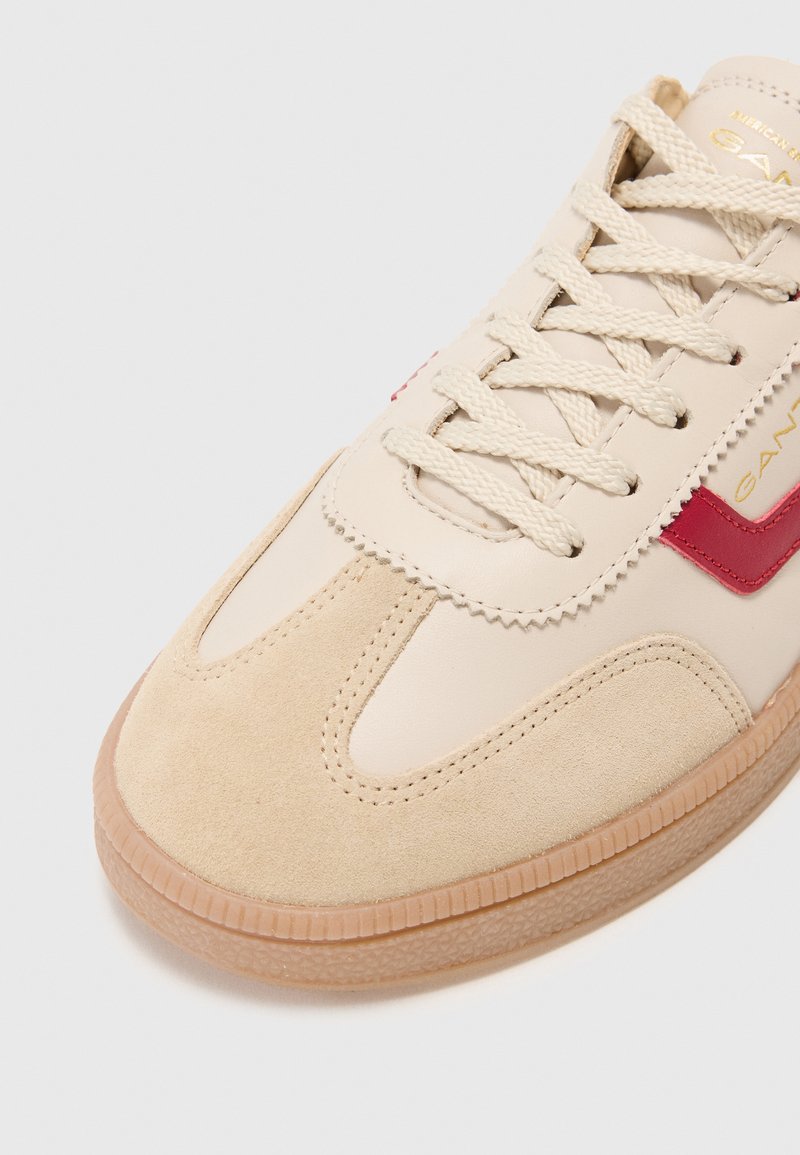 Baskets beige en matières suédées et cuir, avec une bande rouge en accent, une semelle en caoutchouc texturée et des lacets plats beige.