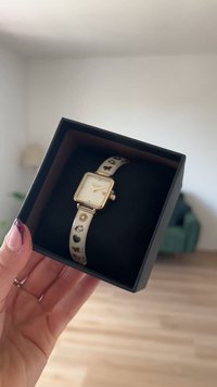 Relógio quadrado dourado com uma bracelete de creme que apresenta pequenos padrões de corações e flores, apresentado numa caixa preta com um interior suave.