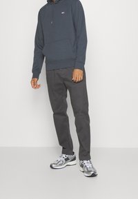 Sudadera gris con bolsillo frontal, pantalones negros ajustados y zapatillas de running plateadas con detalles de malla y acentos negros. Diseño minimalista.