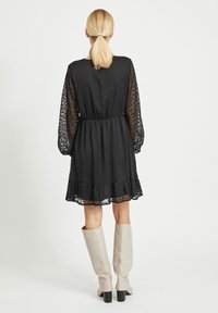 Svart klänning med långa, genomskinliga spetsärmar och en flerlagers volangkant. Kombinerad med knähöga beiga stövlar med klack.