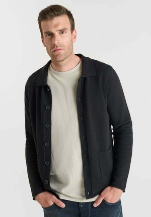 Man in een beige T-shirt en een zwarte knopencardigan, staand met handen in de zakken tegen een effen lichte achtergrond.