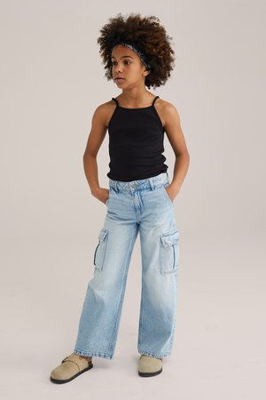 Schwarzes Spaghetti-Träger-Top, hellblaue weit geschnittene Cargo-Jeans mit großen Taschen und beigen Clogs. Strukturiertes lockiges Haar mit einem schwarzen Haarband.