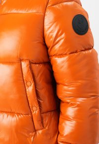 Bright orange puffer jacket featuring a glossy finish, quilted pattern, snap button pocket, and a circular black logo patch on the sleeve.