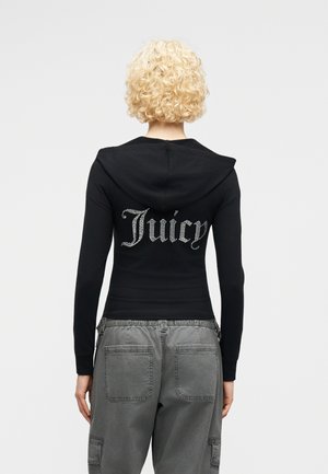 Γυναίκα με κοντά σγουρά ξανθά μαλλιά φοράει μαύρο φούτερ με κουκούλα και το κείμενο "Juicy" από στρας στην πλάτη, καθώς και φαρδιά γκρι παντελόνια cargo.