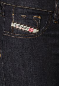 Ciemnoniebieskie dżinsy z małą kieszonką z przodu. Posiadają markową naszywkę z napisem "Diesel Industry" w kontrastowym kolorze oraz kontrastowe przeszycia.