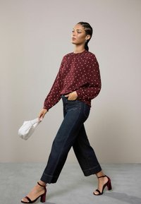 Blusa de lunares burdeos con mangas amplias, combinada con jeans anchos de un azul oscuro. Modelo sosteniendo un bolso blanco acolchado; lleva puestos tacones negros.