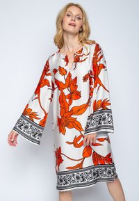 Kwiatowa sukienka tunikowa w białym materiale, z dużymi pomarańczowymi kwiatami, czarnymi akcentami przy rękawach i u dołu oraz dekoltem w serek.