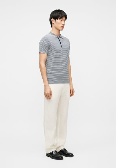 Homme debout portant un polo à manches courtes rayé bleu marine et blanc, un pantalon blanc et des chaussures habillées noires, sur un fond blanc uni.