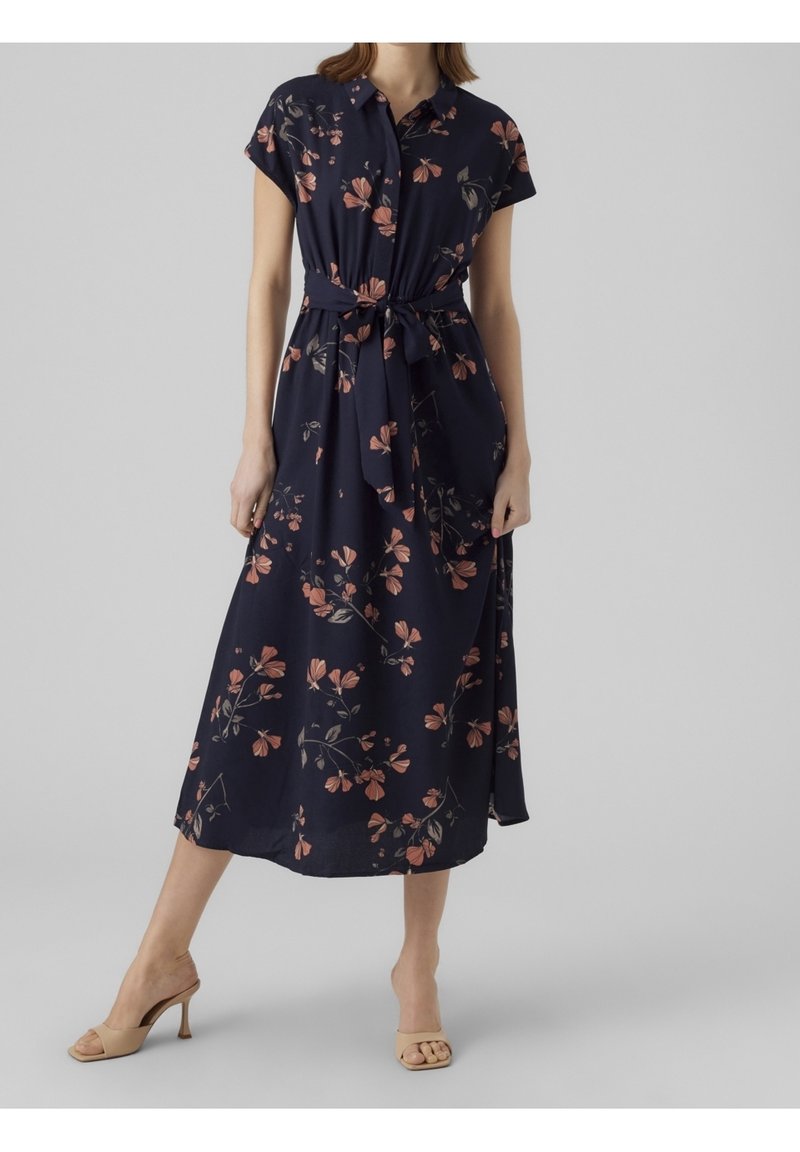 Robe midi bleu marine à manches courtes, motif floral rose, boutonnée sur le devant, taille nouée, portée avec des sandales à talons ouvertes beige.