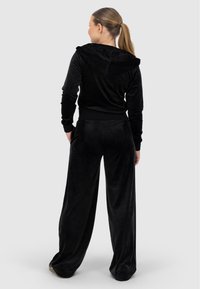 Melna velūra sporta tērps ar pieguļošu kapuci un platām biksēm. Mīksta tekstūra, elastīgas aproces un josta, divas sānu kabatas uz biksēm.