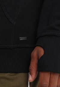 Felpa nera con finitura strutturata, design con scollo a V, polsini a coste e etichetta sul davanti. Include un foro per il pollice in un polsino.