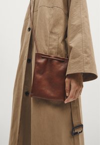 Brązowa skórzana torba na ramię z zaciąganym zapięciem, prostokątny kształt, gładka tekstura, trzymana przy beżowym płaszczu z czarnymi guzikami.